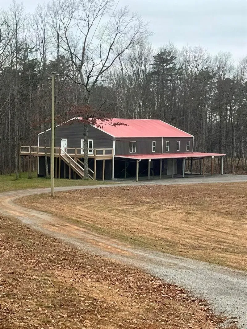 Barndominium with a red roof and wraparound porch beside a gravel driveway